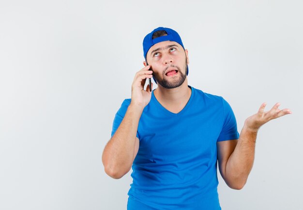 Young man in blue t-shirt and cap talking on mobile phone and looking hesitant