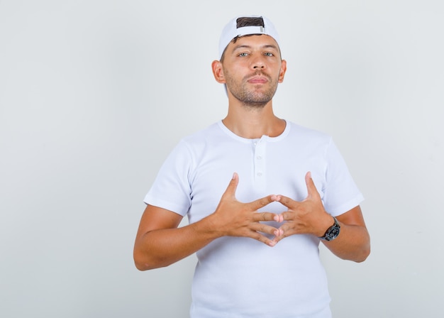 Young man bringing fingers together in white t-shirt, cap and looking proud, front view.