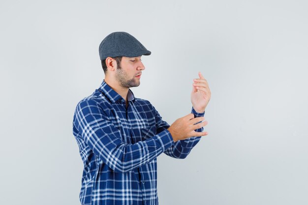 Young man buttoning sleeve of his shirt in shirt, cap and looking handsome , front view.