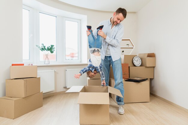 Young man carrying his toddler son upside down under the cardboard box at new home