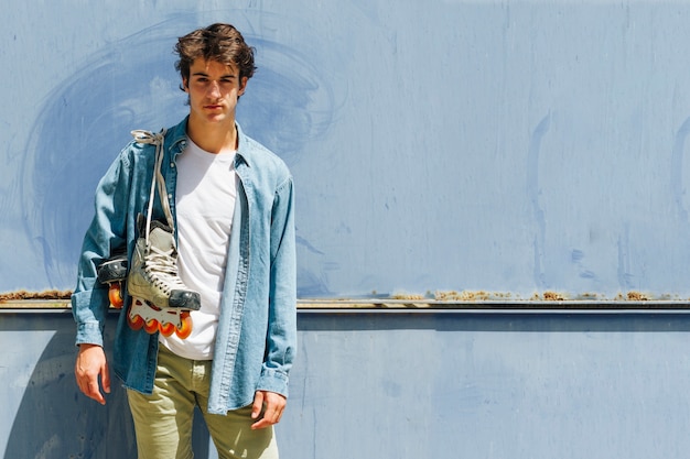 Young man carrying roller skate standing against blue background looking at camera