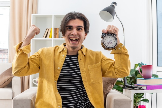 Free photo young man in casual clothes holding alarm clock happy and excited raising fist like a winer sitting on the chair in light living room