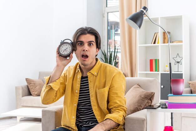 Young man in casual clothes holding alarm clock looking surprised sitting on the chair in light living room