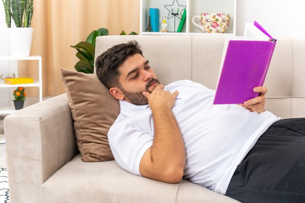 Young man in casual clothes holding book reading with serious face spending weekend at home laying on a couch in light living room