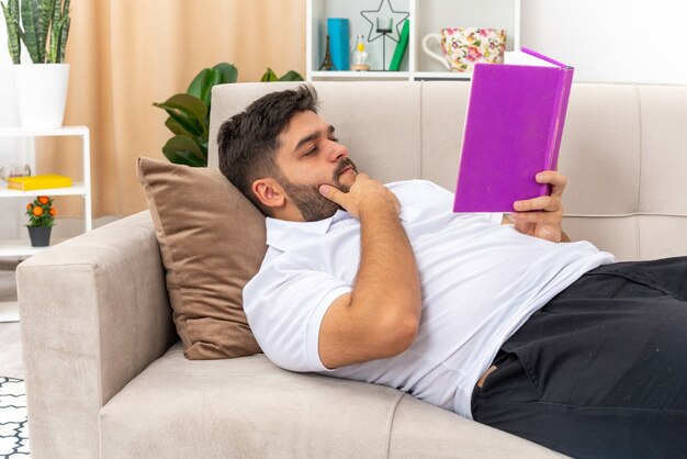 Young man in casual clothes holding book reading with serious face spending weekend at home laying on a couch in light living room