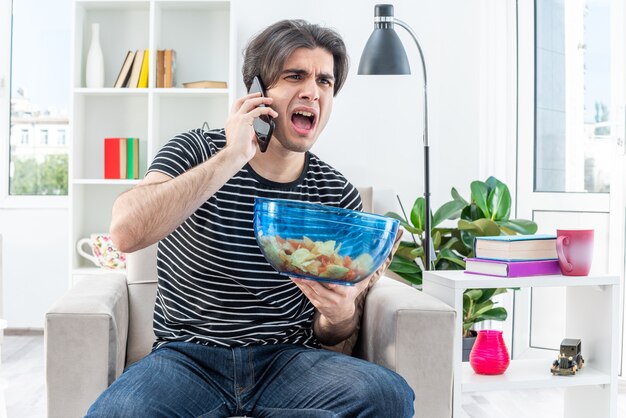 Young man in casual clothes holding bowl of chips shouting being displeased while talking on mobile phone sitting on the chair in light living room