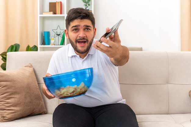 Young man in casual clothes holding bowl of chips and smartphone looking confused sitting on a couch in light living room