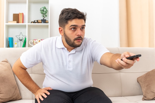 Young man in casual clothes holding tv remote watching tv looking intrigued spending weekend at home sitting on a couch in light living room