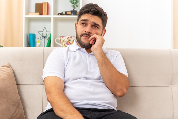 Young man in casual clothes looking aside puzzled sitting on a couch in light living room