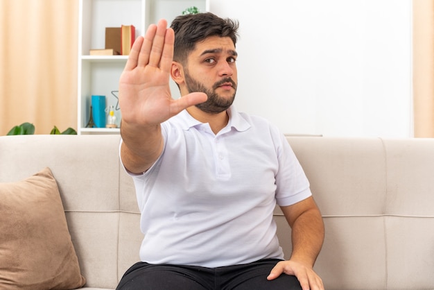 Young man in casual clothes looking with serious face making stop gesture with hand sitting on a couch in light living room