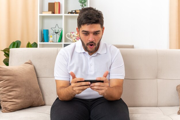 Young man in casual clothes playing games using smartphone emotional and surprised sitting on a couch in light living room