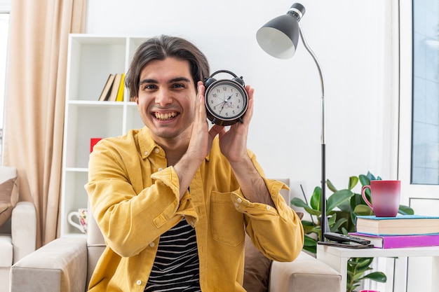 Young man in casual clothes showing alarm clock  happy and cheerful smiling broadly sitting on the chair in light living room