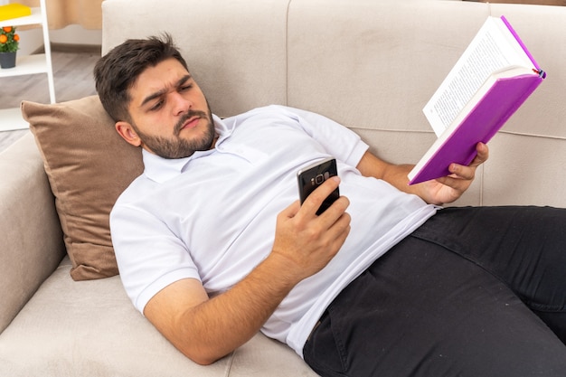 Young man in casual clothes with book holding smartphone looking at it with serious face spending weekend at home laying on a couch in light living room