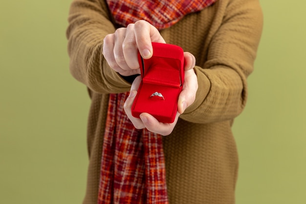 Free photo young man in casual clothes with scarf around neck showing red box with engagement ring valentines day concept standing over green wall