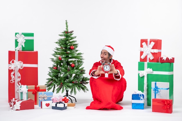 Young man celebrate christmas holiday sitting in the ground and showing clock near gifts and decorated xmas tree