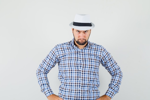 Young man in checked shirt, hat holding hands on waist and looking serious , front view.