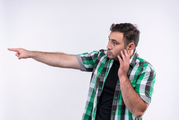 Free photo young man in checked shirt pointing with finger to the side looking confused while talking on mobile phone standing over white wall