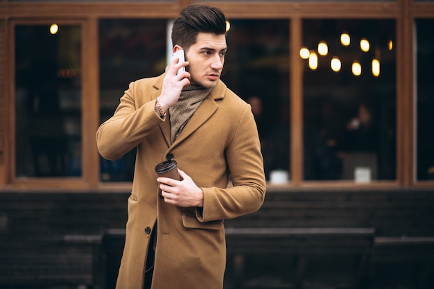 Free photo young man in coat outside talking on the phone and drinking coffee