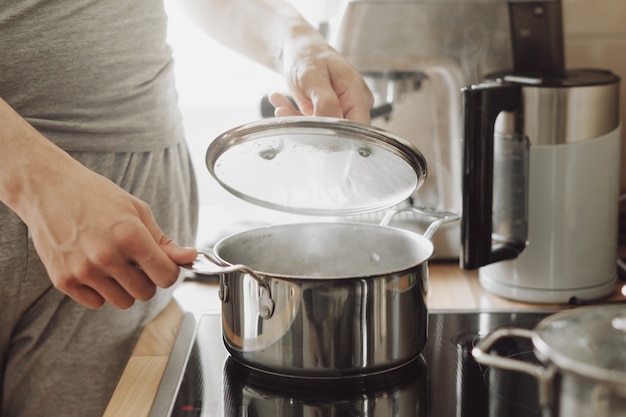 Young man cooking fresh food at home and opening lid of steaming pot.