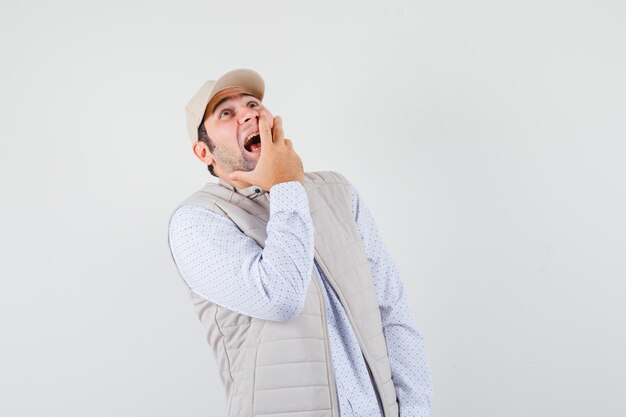 Young man covering mouth with hand, looking upward in beige jacket and cap and looking happy. front view.