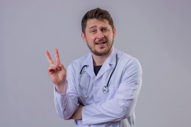 Young man doctor wearing white coat and stethoscope smiling and showing number two or victory sign