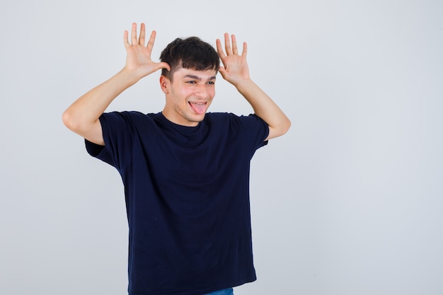 Young man doing funny gesture, sticking out tongue in black t-shirt and looking amused , front view.