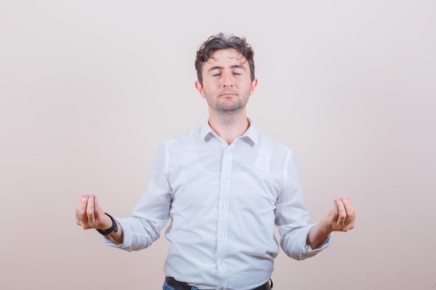 Young man doing meditation with closed eyes in white shirt and looking peaceful