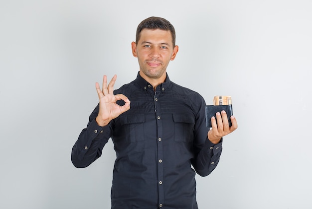 Young man doing ok sign with wallet in black shirt and looking happy