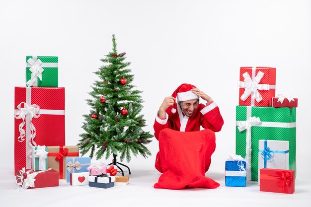 Young man dressed as Santa claus with gifts and decorated Christmas tree sitting in the ground putting both of hands