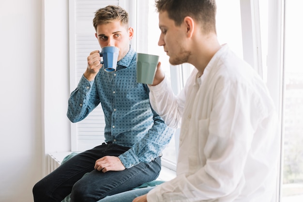 Free photo young man drinking coffee sitting near window