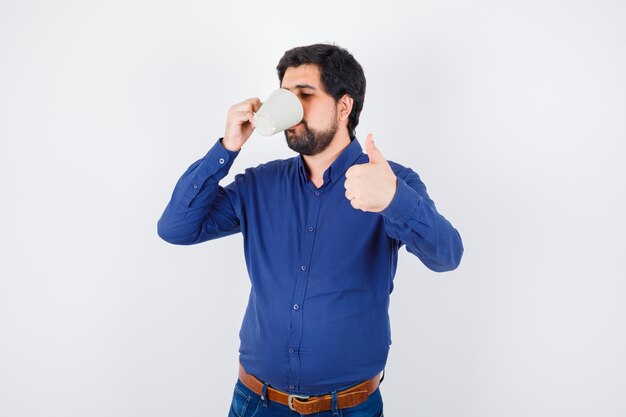 Young man drinking cup of water and showing thumb up in blue shirt and jeans and looking optimistic. front view.