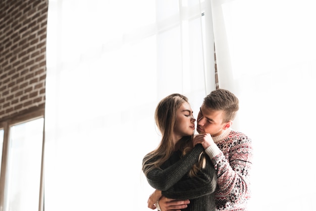 Young man embracing her girlfriend from behind standing against near the window