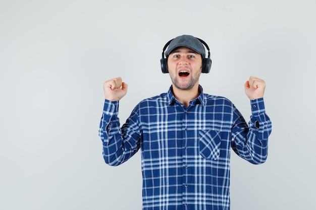 Free photo young man enjoying music with headphones in shirt, cap and looking happy , front view.