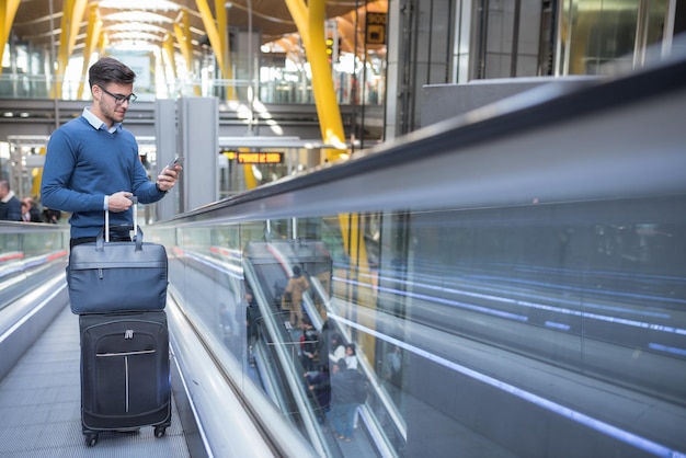 Free photo young man on the escalator at the airport using his mobile phone with his luggage smiling