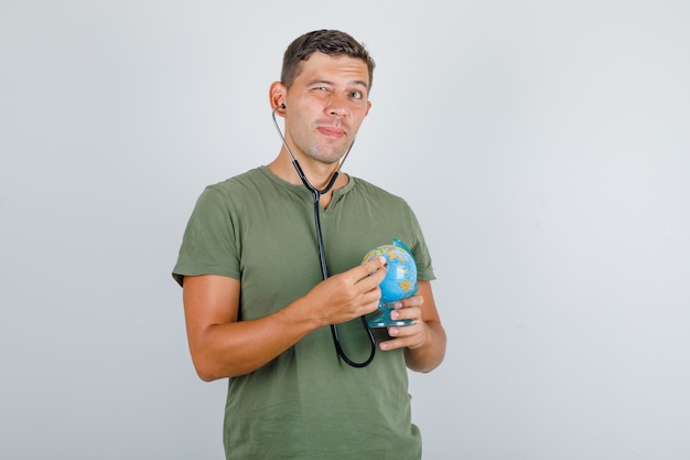 Young man examining globe with stethoscope in army green t-shirt, front view.