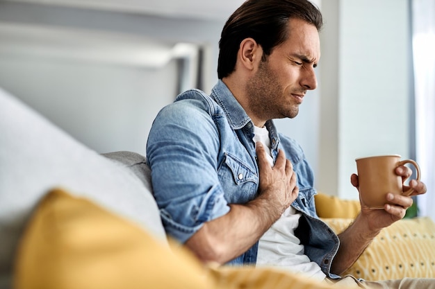 Free photo young man feeling sick and holding his chest in pain while drinking tea in the living room