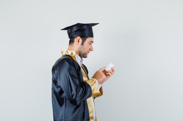 Young man in graduate uniform reading info on bottle of pills and looking curious .