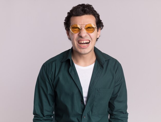 Young man in green shirt wearing glasses annoyed and irritated screaming with closed eyes standing over white wall