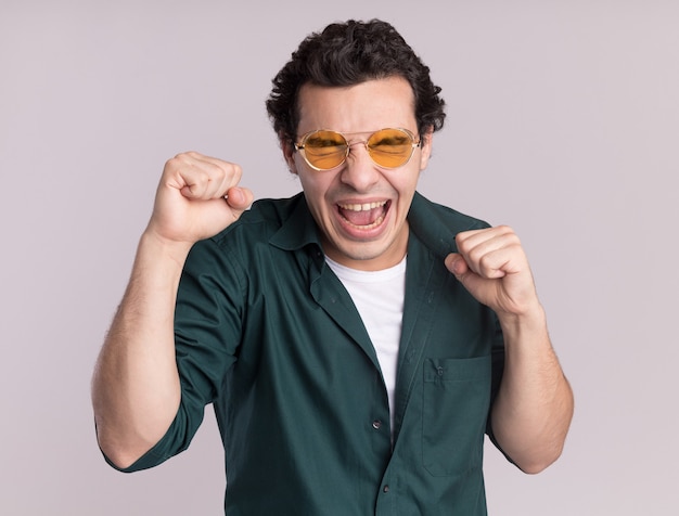 Young man in green shirt wearing glasses happy and excited clenching fists rejoicing his success standing over white wall