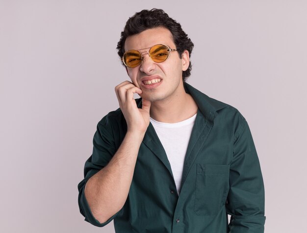 Young man in green shirt wearing glasses looking confused scratching his face standing over white wall