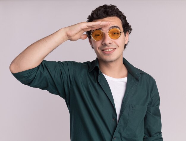 Young man in green shirt wearing glasses looking far away with hand over head standing over white wall