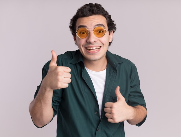 Young man in green shirt wearing glasses looking at front happy and positive smiling cheerfully showing thumbs up standing over white wall