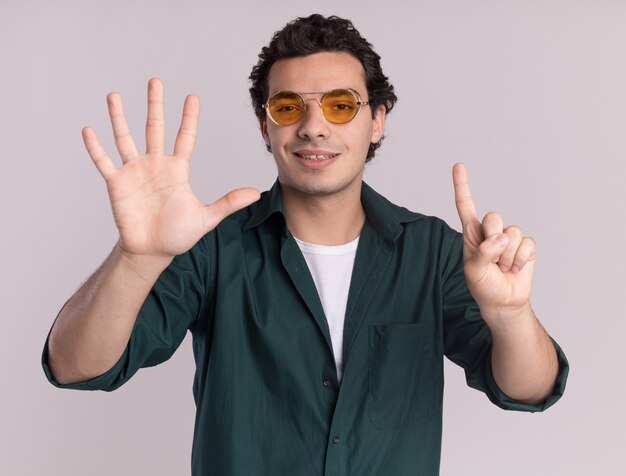 Young man in green shirt wearing glasses looking at front smiling confident showing number six standing over white wall