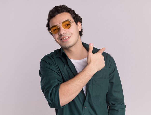 Young man in green shirt wearing glasses looking at front smiling pointing with finger back standing over white wall