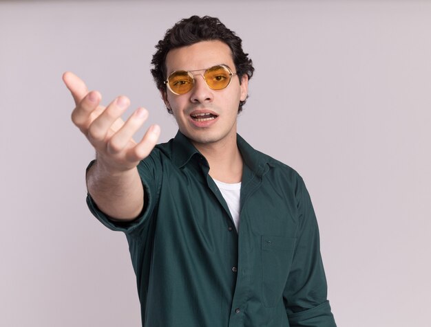 Young man in green shirt wearing glasses looking at front with arm out asking standing over white wall