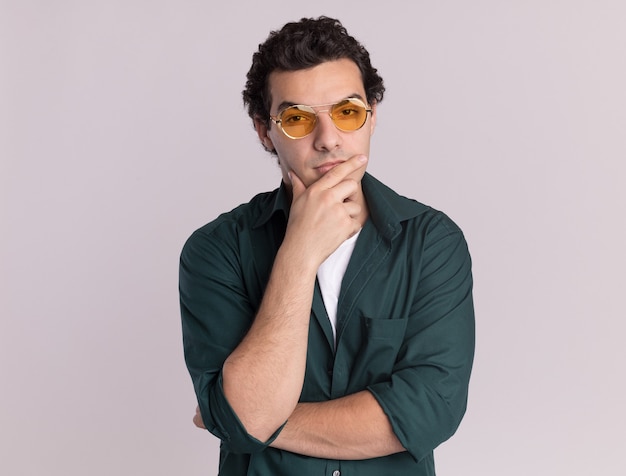 Young man in green shirt wearing glasses looking at front with hand on his chin thinking standing over white wall