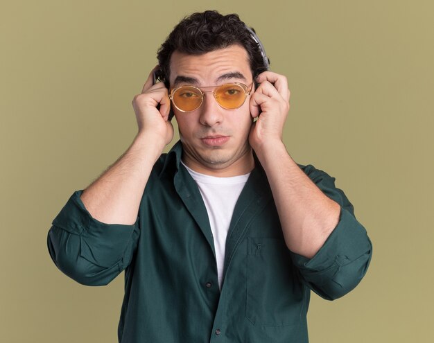 Young man in green shirt wearing glasses with headphones looking at front confused standing over green wall