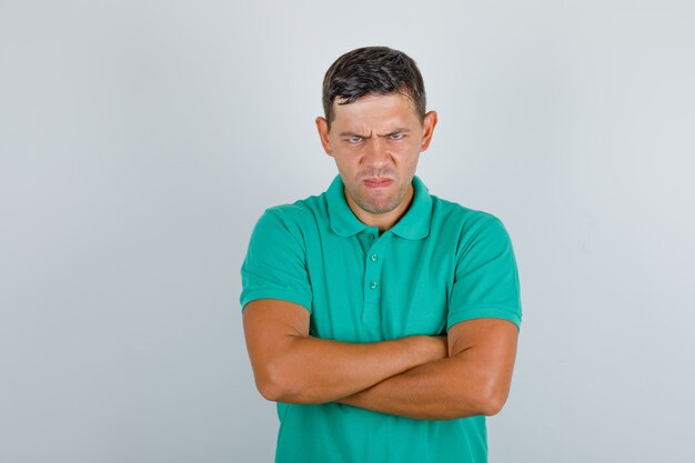 Young man in green t-shirt standing with crossed arms and looking angry, front view.