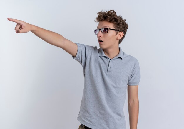 Young man in grey polo shirt looking surprised pointing with index finger at something standing over white wall