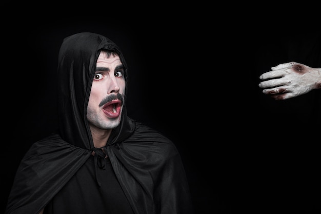 Young man in Halloween costume with scared face and hand of corpse